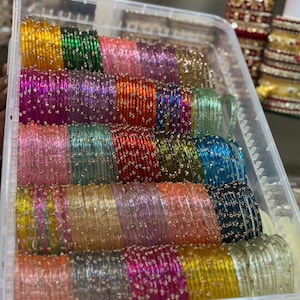 Puede incluir: Un contenedor de almacenamiento de plástico transparente lleno de filas de brazaletes de vidrio multicolores. Cada brazalete presenta un acabado translúcido con pequeños detalles de purpurina plateada incrustados. El texto ZiyaFab es visible arriba a la derecha.