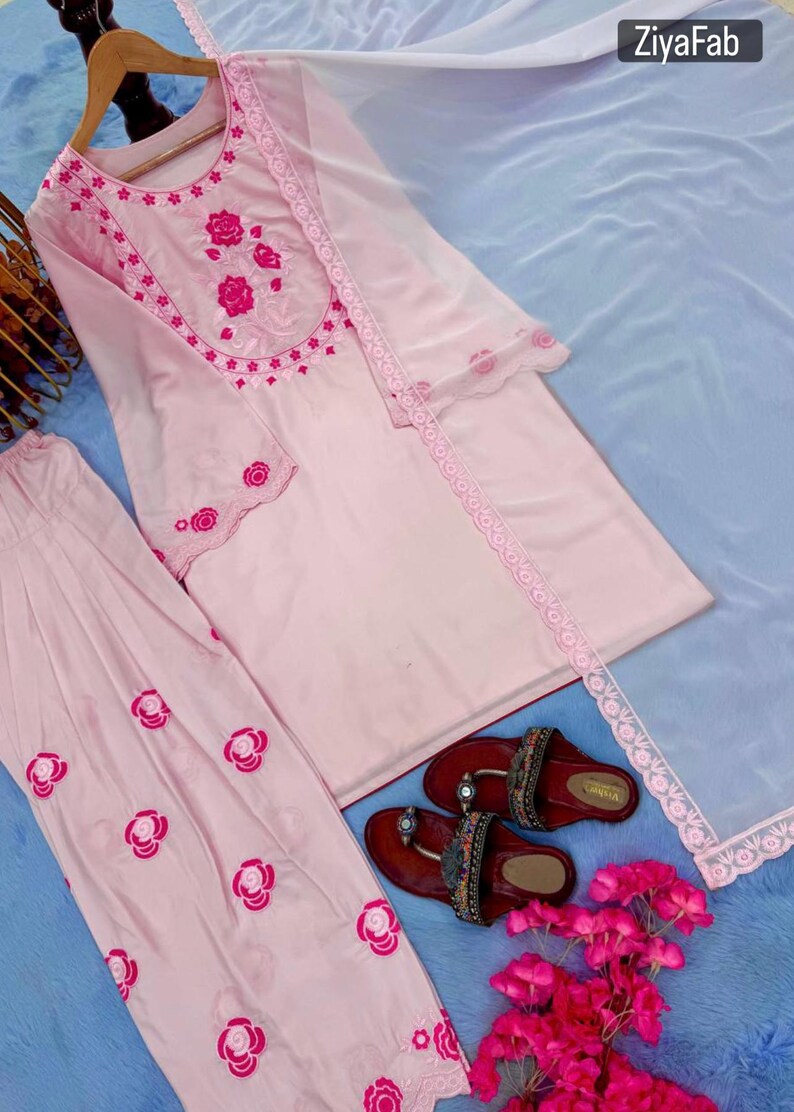 May include: A light pink embroidered salwar kameez set with a matching dupatta. The set features floral embroidery and lace trim. Brown sandals with embellishments are also visible.