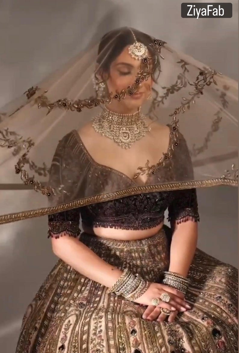 May include: An ornate, gold-toned bridal ensemble. The outfit includes a heavily embellished crop top, a full skirt, and a sheer veil with decorative trim. The model is wearing a matching necklace, bracelets, and rings. The brand name "ZiyaFab" is visible.