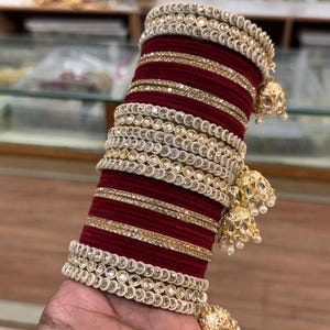 Pode incluir: Um conjunto de pulseiras bordô e douradas com detalhes intrincados. As pulseiras apresentam fileiras de pequenas pedras brilhantes e detalhes dourados. Pingentes dourados pendurados com pérolas adicionam um toque decorativo. O texto "ZiyaFab" é visível.