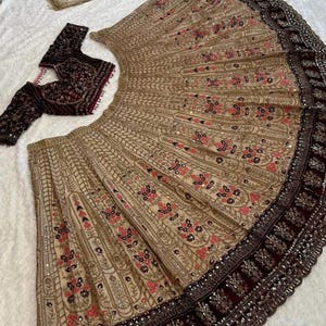 May include: A beige and maroon embroidered lehenga set. The lehenga features intricate floral patterns with gold and pink accents. A matching maroon blouse and a sheer dupatta with floral embroidery complete the ensemble. The brand name "ZiyaFab" is visible.
