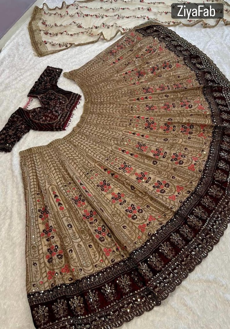 May include: A beige and maroon embroidered lehenga set. The lehenga features intricate floral patterns with gold and pink accents. A matching maroon blouse and a sheer dupatta with floral embroidery complete the ensemble. The brand name "ZiyaFab" is visible.