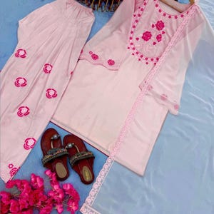 May include: A light pink embroidered salwar kameez set with matching pants and a sheer dupatta. The set features floral embroidery and lace trim. A pair of maroon sandals with decorative embellishments are also present.