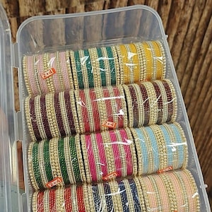 May include: A clear plastic storage box containing twelve sets of stacked bangles in various colors including pink, green, yellow, maroon, blue, and red. Each set features alternating metallic gold-toned beads and colored bands with 2x4 labels.