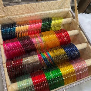 Può includere: Una collezione di braccialetti in vetro traslucido e colorato disposti in un portagioie beige con il testo ZiyaFab visibile. I braccialetti presentano una varietà di tonalità vivaci tra cui rosso, verde, giallo, blu, rosa e toni trasparenti.