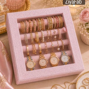 May include: Pink jewellery box with a clear lid, filled with gold and pearl bracelets, bangles, and four gold watches. The watches have gold and silver bands and cream-coloured faces. The box has the text "ZiyaFab".