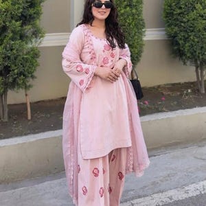 May include: A light pink salwar kameez set with floral embroidery. The outfit includes a tunic, pants, and a matching scarf. The tunic and pants feature a repeating floral pattern. The outfit is worn with sunglasses and a black purse. The text "ZiyaFab" is in the top right corner.