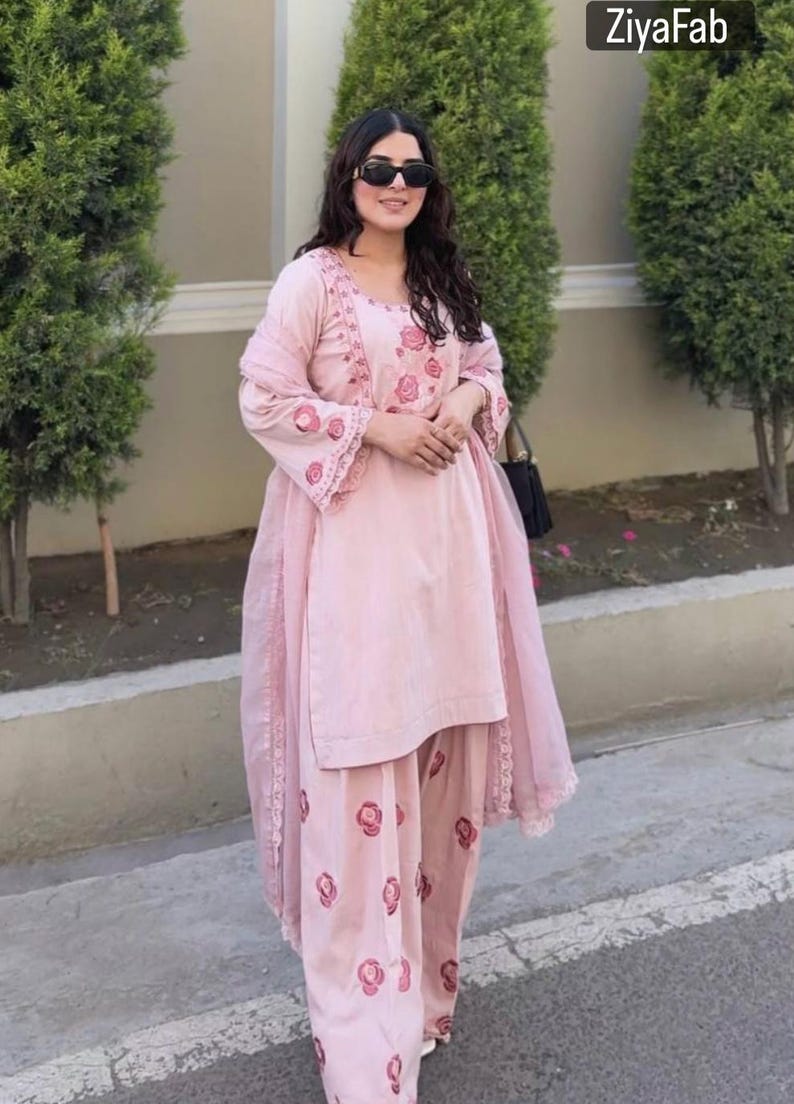 May include: A light pink salwar kameez set with floral embroidery. The outfit includes a tunic, pants, and a matching scarf. The tunic and pants feature a repeating floral pattern. The outfit is worn with sunglasses and a black purse. The text "ZiyaFab" is in the top right corner.