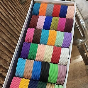 May include: A rectangular white box containing rows of thin, matte-finish metal bangles in a wide variety of solid colors including blue, orange, gray, red, pink, green, yellow, black, white, purple, and gold. The text ZiyaFab is visible.