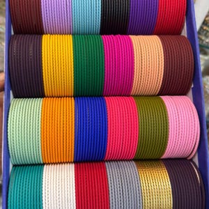May include: A collection of thin metal bangles arranged in a blue box, featuring a wide variety of solid colors including red, yellow, green, blue, purple, pink, orange, white, gray, black, and gold. The text ZiyaFab is visible.