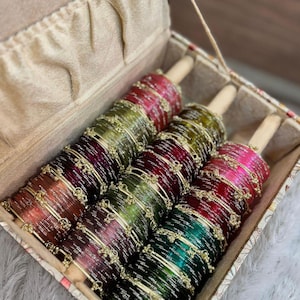 Pode incluir: Uma coleção de pulseiras coloridas numa caixa decorativa. As pulseiras são em tons de vermelho, verde e dourado, e estão dispostas em cavilhas de madeira. A caixa é bege com um padrão floral. O texto "ZiyaFab" está no canto superior direito.