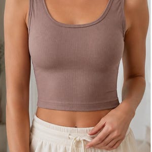 Puede incluir: Un top corto sin mangas de punto acanalado en color malva, con un amplio escote redondo y tirantes anchos. La prenda es ajustada, termina justo por encima de la cintura y se combina con pantalones de chándal de color blanco roto.