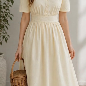 May include: A cream-colored midi dress with short sleeves, a V-neckline, and a button-down front. The dress features a fitted waist and a flowing skirt. A small woven basket is held in the hand.