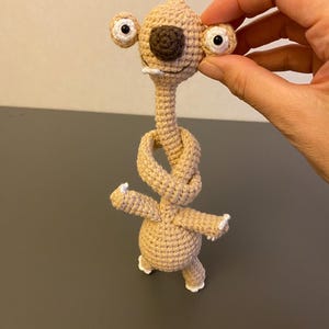 Pode incluir: Um personagem de crochê de cor bege com olhos grandes e redondos, um nariz castanho e um pescoço atado. O personagem tem braços e pernas pequenos com detalhes brancos. O brinquedo de pelúcia feito à mão é segurado por uma mão.