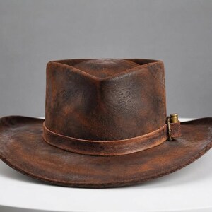 May include: A distressed brown leather cowboy hat with a wide, curved brim and a pinched crown. The hat features a matching leather band around the base, secured with a small, metallic brass-coloured buckle detail on the side of the crown.