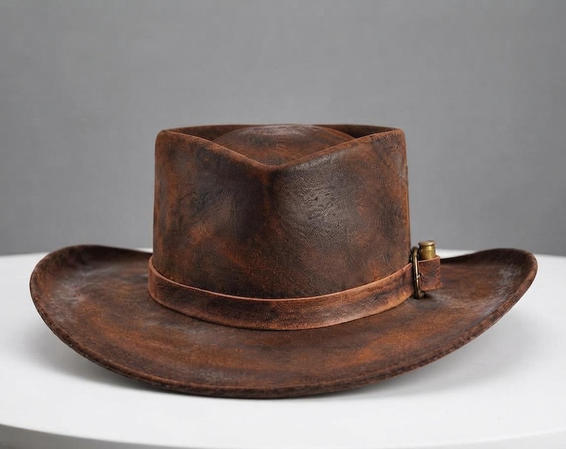 May include: A distressed brown leather cowboy hat with a wide, curved brim and a pinched crown. The hat features a matching leather band around the base, secured with a small, metallic brass-coloured buckle detail on the side of the crown.