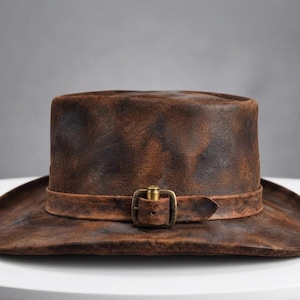 May include: A distressed brown leather hat with a textured, weathered finish. The hat features a matching leather band around the crown, secured by a rectangular brass-toned metal buckle with a central pin, creating a rugged appearance.