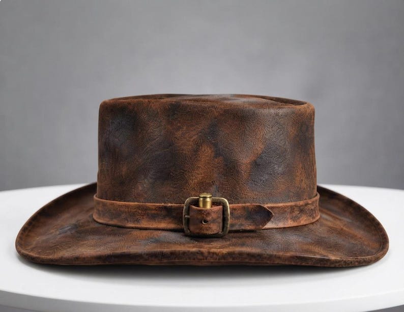 May include: A distressed brown leather hat with a textured, weathered finish. The hat features a matching leather band around the crown, secured by a rectangular brass-toned metal buckle with a central pin, creating a rugged appearance.