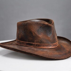 May include: A brown fedora hat with a distressed, weathered texture and a dark, uneven finish. The hat features a pinched crown, a wide brim, and a matching brown band encircling the base of the crown, creating a rugged and worn appearance.