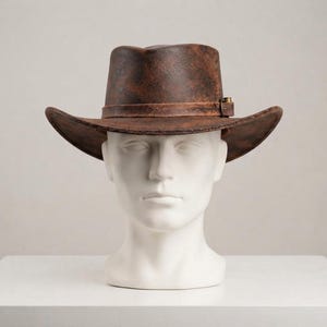 May include: A brown fedora hat with a distressed, weathered texture and a dark brown leather band featuring a small metallic buckle detail. The hat has a structured crown and a wide, slightly curved brim, displayed on a white mannequin head.