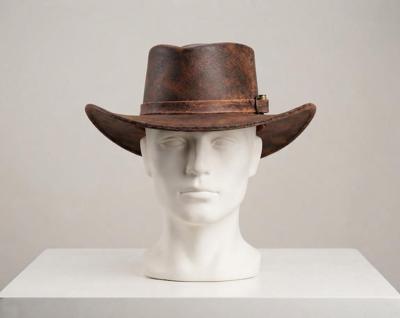 May include: A brown fedora hat with a distressed, weathered texture and a dark brown leather band featuring a small metallic buckle detail. The hat has a structured crown and a wide, slightly curved brim, displayed on a white mannequin head.