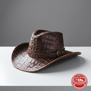 May include: A dark brown cowboy hat featuring a textured crocodile skin pattern across the entire surface. The hat includes a matching brown band around the crown, accented with a small, metallic buckle detail on the side of the brim.