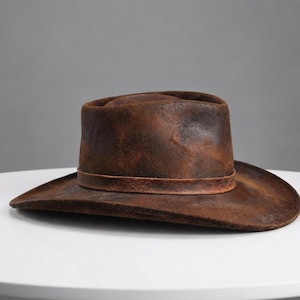 May include: A distressed brown leather cowboy hat with a weathered texture and a matching leather band around the crown. The hat features a structured brim and a deep, rounded crown, showcasing a dark, uneven finish across the entire surface.