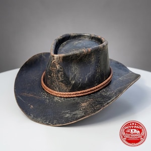 Op de afbeelding: Een verweerde zwarte cowboyhoed met een versleten afwerking die bruine ondertonen laat zien. De hoed heeft een hoge kroon en een brede, gebogen rand. Een dubbel gevlochten bruin koord wikkelt zich als decoratieve band rond de basis van de kroon.