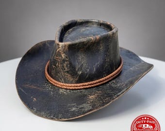May include: A distressed black cowboy hat with a weathered finish and a textured appearance. The hat features a pinched crown and a wide, curved brim. A double-strand braided brown leather cord wraps around the base of the crown as a decorative band.