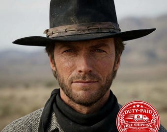 May include: A black felt cowboy hat with a wide brim and a brown leather hat band featuring decorative stitching. The hat has a structured crown and a weathered appearance, paired with a black neckerchief and a textured grey collared shirt.