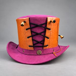 Puede incluir: Un sombrero de copa magenta con un panel de aspecto de cuero color canela y cordones negros cruzados. El sombrero incluye detalles metálicos en tono latón, como pequeños dijes en forma de campana y un botón decorativo circular.