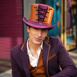 Può includere: Un cappello a cilindro con tesa e base della corona viola, accentuato da un pannello color cuoio sulla parte anteriore. Il pannello è fissato con lacci neri incrociati e occhielli metallici. Un piccolo emblema metallico circolare è centrato sulla fascia.