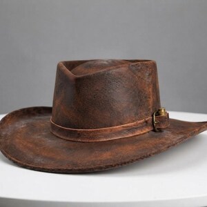 May include: A dark brown, distressed leather cowboy hat with a wide, curved brim and a high crown. A thin leather band wraps around the base of the crown, secured by a small, metallic brass-coloured buckle and a single decorative bullet casing.