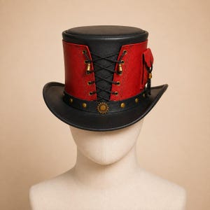 Puede incluir: Un sombrero de copa negro y rojo con un panel central de cordones negros con ojales de latón, un pequeño bolsillo lateral y un ala negra adornada con una banda de tachuelas de latón y un emblema en forma de engranaje en la parte frontal.