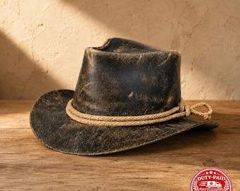 May include: A black cowboy hat with a distressed, weathered finish and a light tan rope hatband wrapped twice around the crown, featuring a knotted detail on the side. The hat has a wide, curved brim and a pinched crown design.