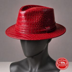 Könnte beinhalten: Ein leuchtend roter Fedora-Hut mit einer strukturierten Krokodilleder-Optik auf der gesamten Krone und Krempe. Der Hut verfügt über ein passendes rotes Band, das um die Basis der Krone gewickelt ist und ein strukturiertes, einheitliches Aussehen bietet.