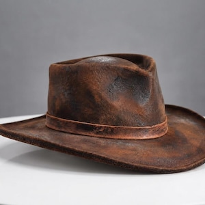 May include: A brown fedora hat with a distressed, weathered texture and a dark, uneven finish. The hat features a pinched crown, a wide brim, and a matching brown band encircling the base of the crown, creating a rugged and worn appearance.