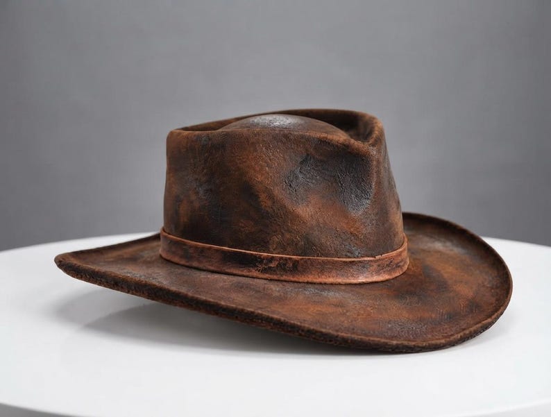 May include: A brown fedora hat with a distressed, weathered texture and a dark, uneven finish. The hat features a pinched crown, a wide brim, and a matching brown band encircling the base of the crown, creating a rugged and worn appearance.