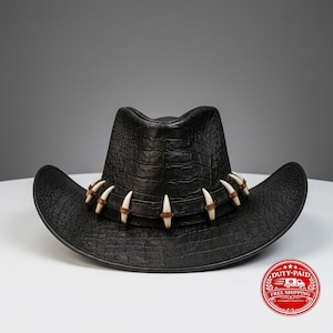 Puede incluir: Un sombrero de vaquero negro con un patrón de textura de piel de cocodrilo. El sombrero está adornado con una banda de cuero negro que rodea la corona, decorada con una serie de acentos curvos de color blanco hueso en forma de dientes.