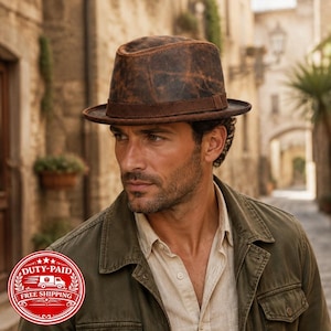 Puede incluir: Un sombrero fedora de cuero marrón desgastado con una banda de ante marrón oscuro. El sombrero presenta una corona pellizcada y un ala estructurada, mostrando una textura envejecida con marcas de color marrón oscuro en la superficie.