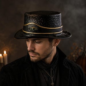 Puede incluir: Un sombrero de copa negro con una superficie texturizada similar a la piel de reptil. El sombrero presenta una banda negra ancha y lisa alrededor de la copa, acentuada por dos finas líneas horizontales doradas y un emblema central de un ojo verde.