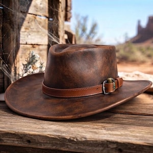 May include: A distressed brown leather cowboy hat with a wide brim and a pinched crown. The hat features a smooth, thin brown leather band wrapped around the base, secured by a metallic buckle with a small brass-coloured bullet casing detail.