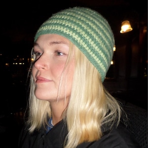 May include: A green and light green striped crochet beanie. The hat is worn by a person with blonde hair and a black jacket. The background is dark.