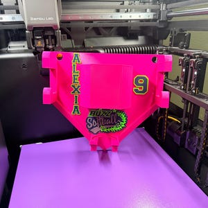 Peut inclure: Un masque de receveur de softball rose vif avec le nom "ALEXIA" en lettres vertes. Le numéro "9" est également en vert. Le masque porte les mots "BUZZ Softball" dans un design vert et noir. Le masque est en cours d'impression 3D.