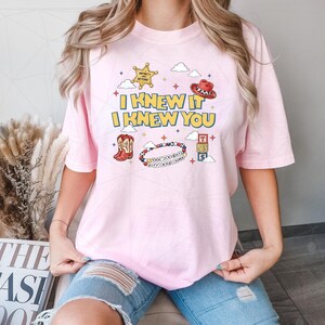 May include: A light pink t-shirt featuring a graphic print with the text I KNEW IT I KNEW YOU, a yellow sheriff star, a red cowboy hat, red cowboy boots, a colorful beaded bracelet with text, and a stack of three lettered blocks with clouds.
