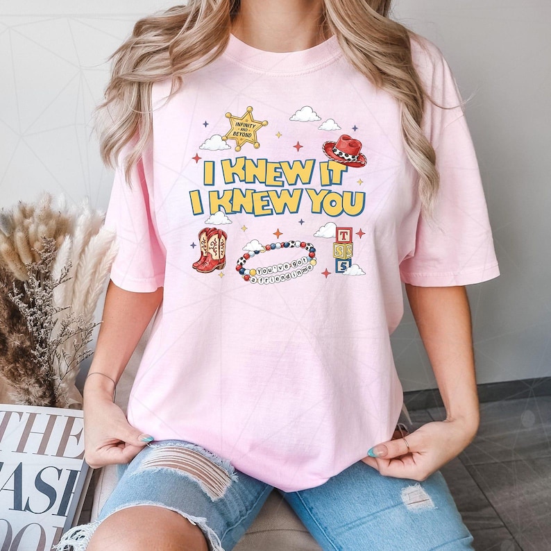 May include: A light pink t-shirt featuring a graphic print with the text I KNEW IT I KNEW YOU, a yellow sheriff star, a red cowboy hat, red cowboy boots, a colorful beaded bracelet with text, and a stack of three lettered blocks with clouds.