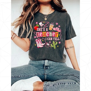 May include: A charcoal gray short-sleeve t-shirt featuring a colorful graphic print with the text "SHE'S A teacher I CAN TELL". The design includes illustrations of a red apple, laptop, pencils, iced coffee, pink cowboy hat, boots, and a cactus.