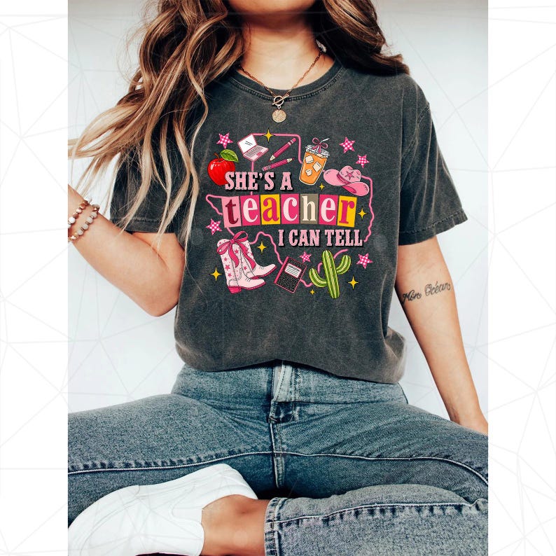 May include: A charcoal gray short-sleeve t-shirt featuring a colorful graphic print with the text "SHE'S A teacher I CAN TELL". The design includes illustrations of a red apple, laptop, pencils, iced coffee, pink cowboy hat, boots, and a cactus.