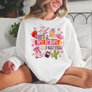 May include: A white crewneck sweatshirt featuring a colorful graphic print with the text "SHE'S A TEACHER I CAN TELL". The design includes illustrations of a red apple, a laptop, pencils, an iced coffee, a pink cowboy hat, boots, and a cactus.