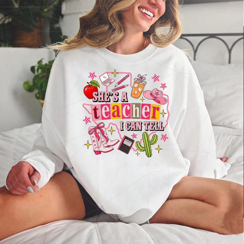 May include: A white crewneck sweatshirt featuring a colorful graphic print with the text "SHE'S A TEACHER I CAN TELL". The design includes illustrations of a red apple, a laptop, pencils, an iced coffee, a pink cowboy hat, boots, and a cactus.